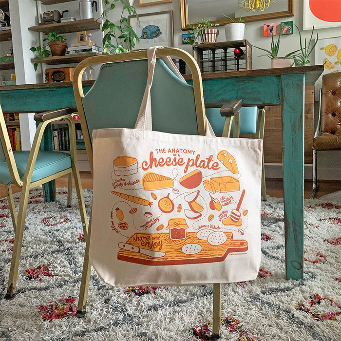 In a colorful dining room, a teal chair and wooden table set the scene. Hanging on the chair is The Neighborgoods Tote Bag, adorned with cheese illustrations and labeled The Anatomy of a Cheese Plate. This eco-friendly bag adds charm amid the plants and decor in the background.
