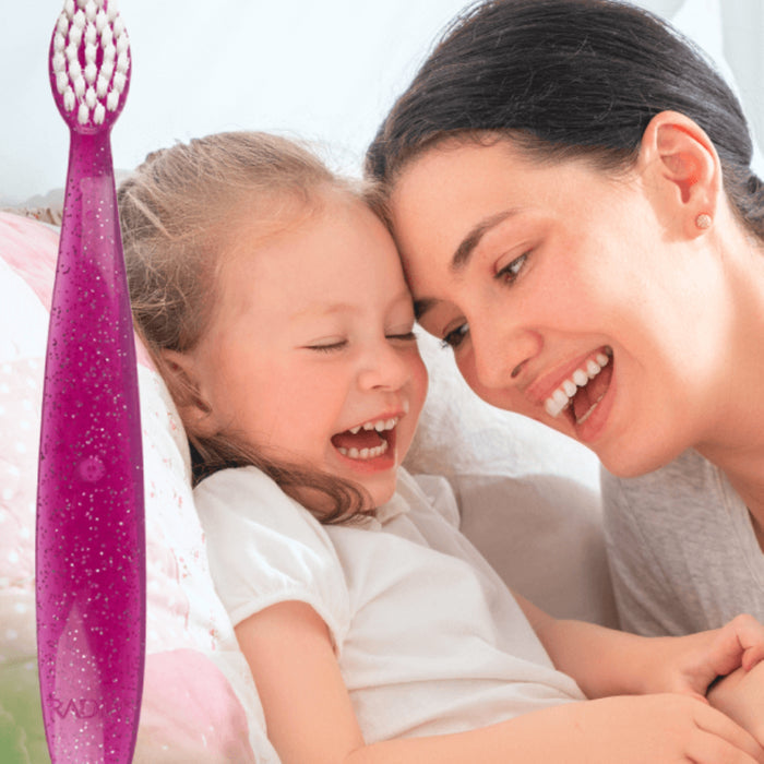A smiling woman and a young girl laugh together on a bed beside a Radius Totz Toothbrush 18 mo+, capturing a joyful and playful moment.