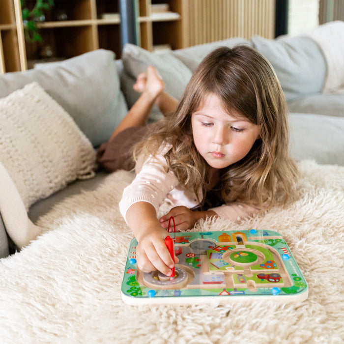A young child with long hair lies on a fluffy cream rug, focused on the colorful Town Maze Magnetic Puzzle Game by HABA. Using a red stylus, they navigate this puzzle, honing fine motor skills, while a couch and shelves remain in the background.
