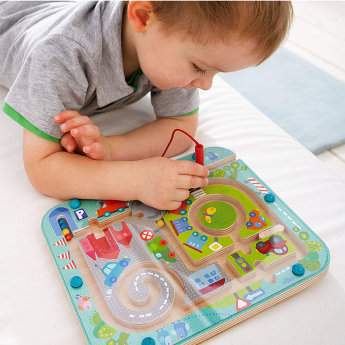 A young child boosts fine motor skills with HABAs Town Maze Magnetic Puzzle Game, featuring roads, cars, and a roundabout. Using the red magnetic pen, they guide small beads through the maze on a light surface.