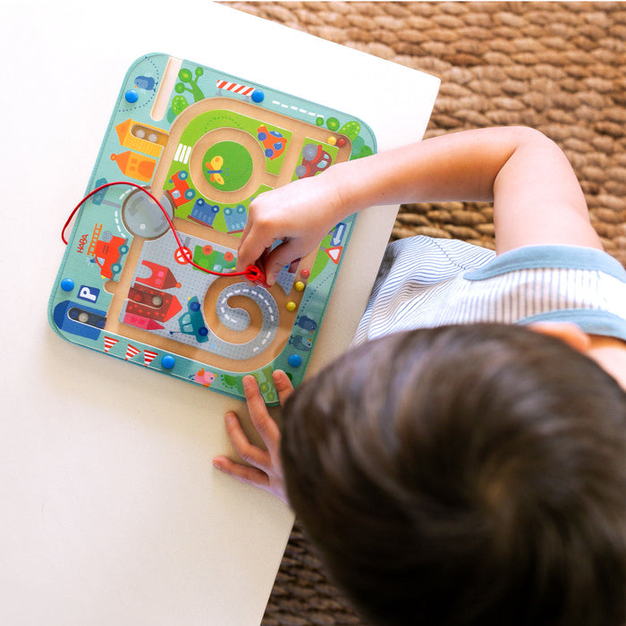 A child is captivated by the Town Maze Magnetic Puzzle Game from HABA, navigating beads through a vibrant maze of roads, buildings, and a firefighter illustration using a magnet wand at a white table on a textured brown surface, enhancing fine motor skills.