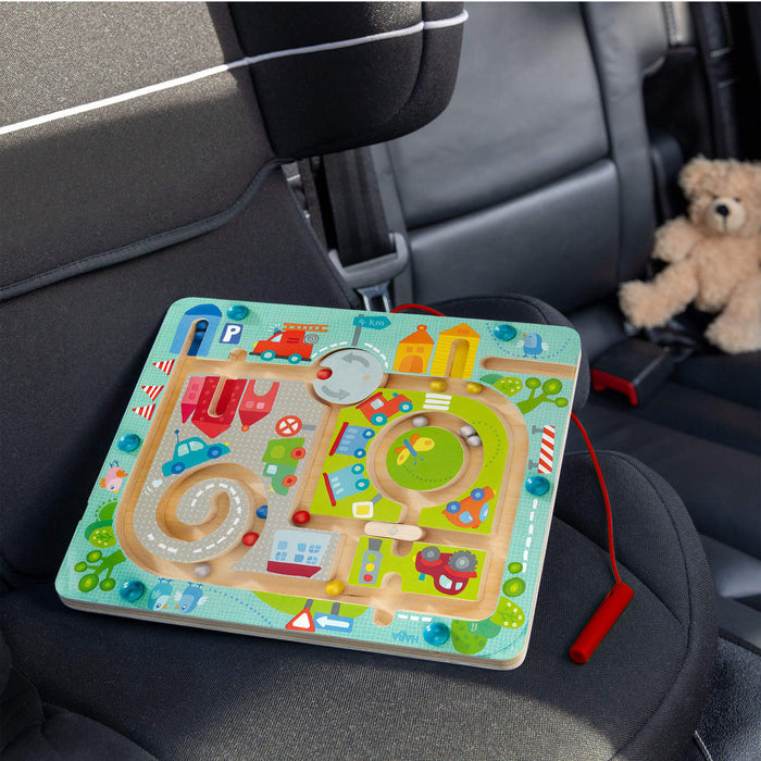 The Town Maze Magnetic Puzzle Game by HABA sits on a car seat. This colorful childrens toy features roads, buildings, and vehicles navigated by a magnetic wand, perfect for enhancing fine motor skills. A teddy bear watches over the fun scene in the background.