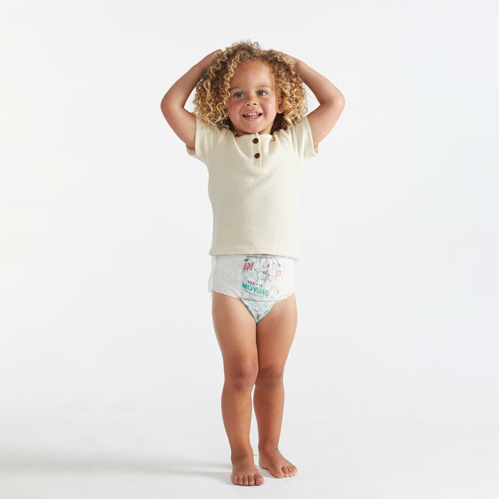 A curly-haired child smiles, hands on head, dressed in a light shirt and The Honest Companys Training Pants - 3T/4T, featuring an absorbent core, against a plain white background.