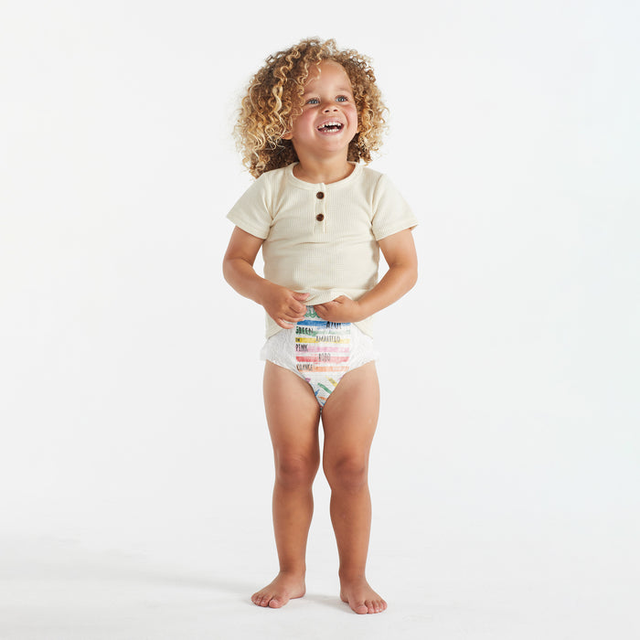 A child with curly hair smiles against a plain background, wearing a light shirt and The Honest Companys colorful Training Pants in size 4T/5T, ideal for potty-training adventures.