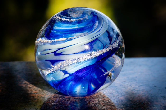 Blue and white glass swirl paperweight with ash specks photographed outdoors on a granite surface