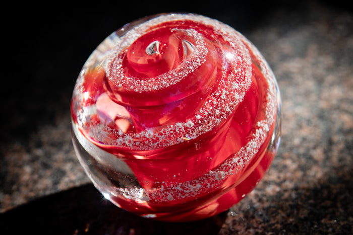 Red swirling glass paperweight with embedded cremation ashes, sunlit on a granite countertop.