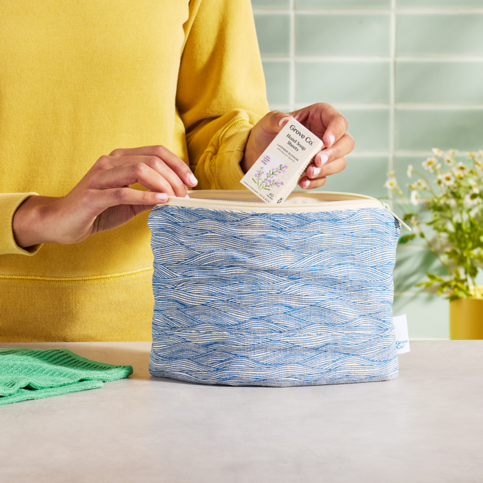 A person in a yellow sweater places a lavender-scented sachet into a Grove Co. Travel Pouch, crafted from 100% organic cotton. A green cloth complements the eco-style vibe, against a backdrop of tiled walls and a vase with flowers.