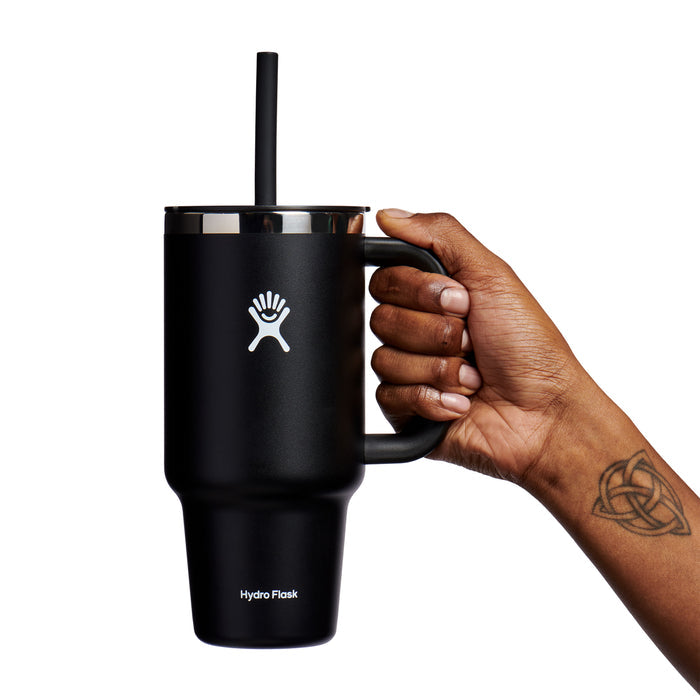A tattooed hand holds a black 32 oz Hydro Flask Travel Tumbler with a straw and built-in handle, elegantly set against a white background. This insulated tumbler features double-wall vacuum insulation for superior temperature retention.