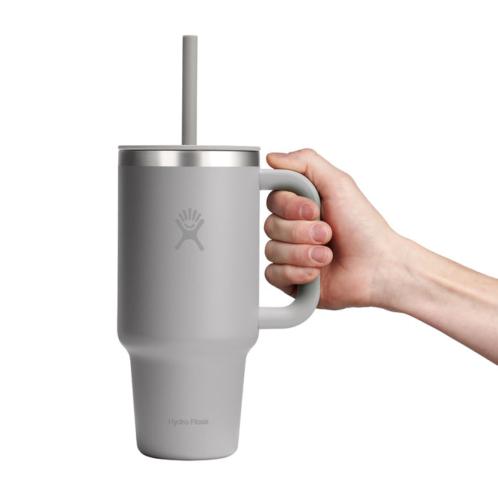 A hand holds a 32 oz gray Hydro Flask Travel Tumbler with a handle and straw. It has a matte finish, features the Hydro Flask logo, and offers double-wall vacuum insulation for optimal temperature retention.