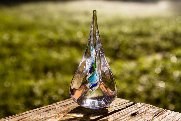 Tri-color glass memorial flame with cremation ash visible in a spiral, displayed outdoors on a wooden surface.
