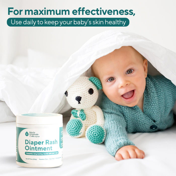 A smiling baby in a teal sweater lies under a white blanket with a crocheted bear and Marie Originals Triple Action Diaper Rash Ointment. Text above recommends daily use of this ointment for healthy, happy skin.