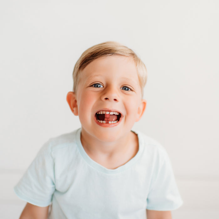A joyful child with light brown hair in a light blue shirt smiles wide, revealing a missing tooth. Their smile is as refreshing as True Graces True Littles Daily Kids Multivitamin on a warm day, with the plain white background highlighting their happiness.