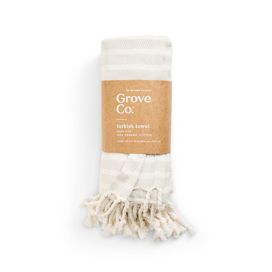 A folded ivory Turkish towel with beige stripes, labeled Grove Co. from the Spring Collection, features an organic cotton tag. It has tasseled edges and is wrapped in a brown eco-friendly paper band emphasizing its sustainable commitment.