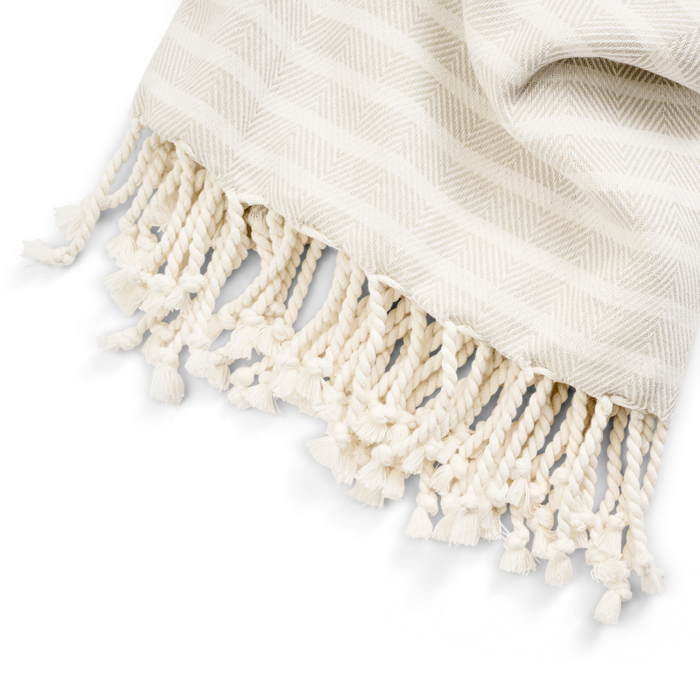 The Large Turkish Towel from Grove Co.s Spring Collection is elegantly displayed with a beige and cream herringbone design. Made from sustainably sourced materials, it boasts twisted fringes for texture. Its soft, cozy fabric ensures comfort and style in every drape.