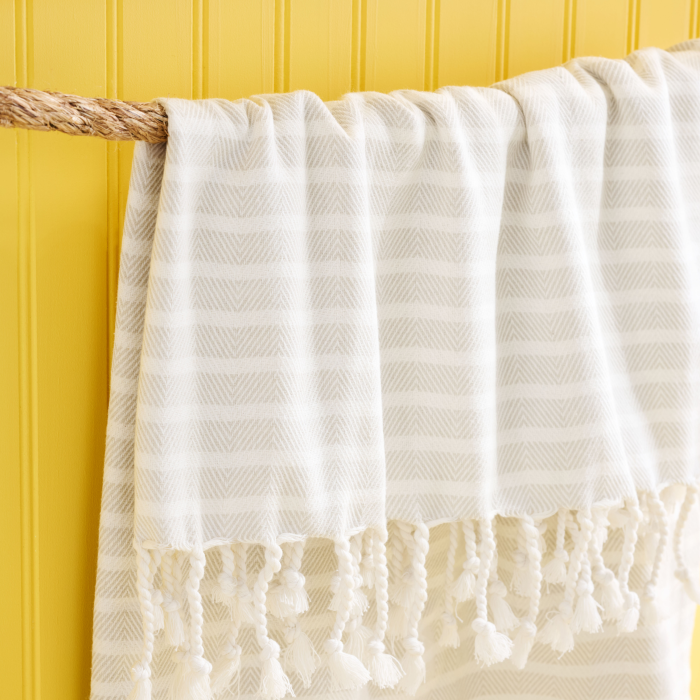 The Large Turkish Towel from Grove Co.s Spring Collection, crafted from organic cotton in cream with a subtle chevron pattern and fringe, hangs elegantly over a thick rope against a bright yellow vertical-panel wall.