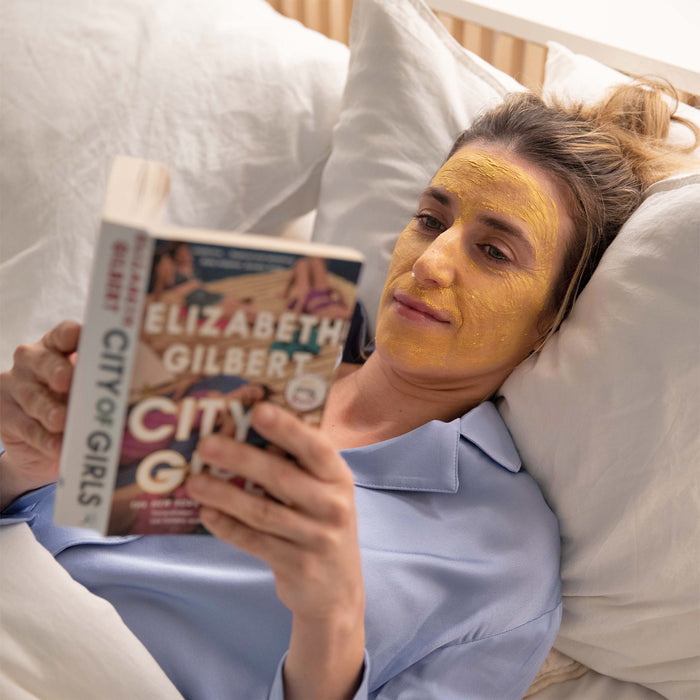 A woman wearing the APTO Skincare Turmeric Mask with Azelaic Acid, Soothing & Clarifying Face Mask relaxes in bed. She reads City of Girls by Elizabeth Gilbert in a light blue pajama top against white pillows, deeply immersed in her book.