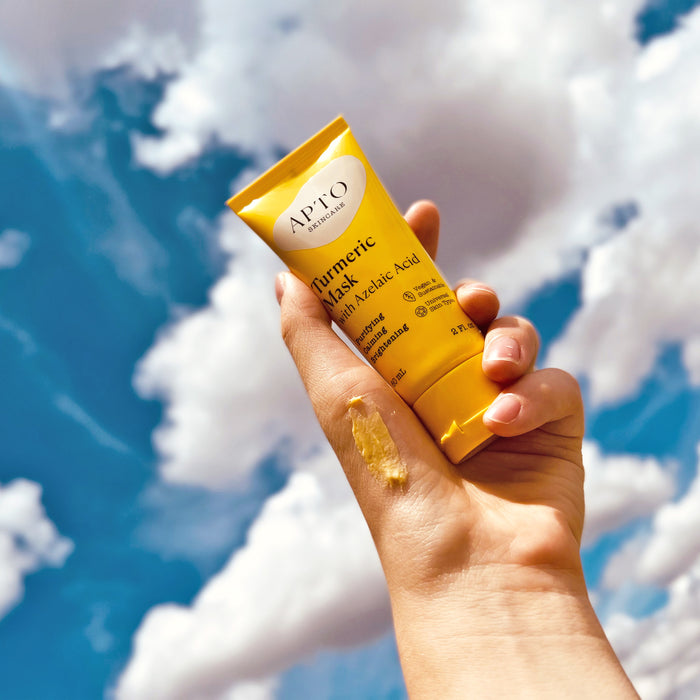 A hand holds a yellow tube of APTO Skincares Turmeric Mask with Azelaic Acid against a blue sky with white clouds, while a small amount is applied on the back of the hand.