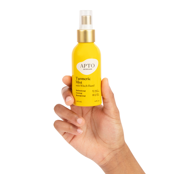 A hand holds APTO Skincares Turmeric Mist with Witch Hazel, a Reviving & Brightening Facial Spritz in a bright yellow bottle with a gold spray cap, promising glowing skin. The cruelty-free face mist displaying APTO Skincare stands out against the plain black background.