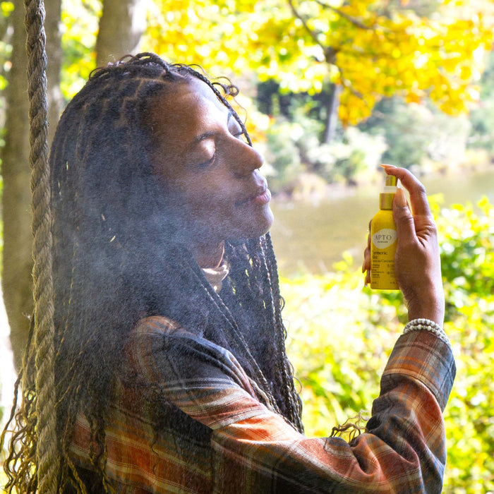 A person with long braided hair enjoys APTO Skincares cruelty-free Turmeric Mist with Witch Hazel on their skin. They are outside in a plaid shirt, looking serene as sunlight filters through green and yellow leaves, enhancing their glowing complexion.