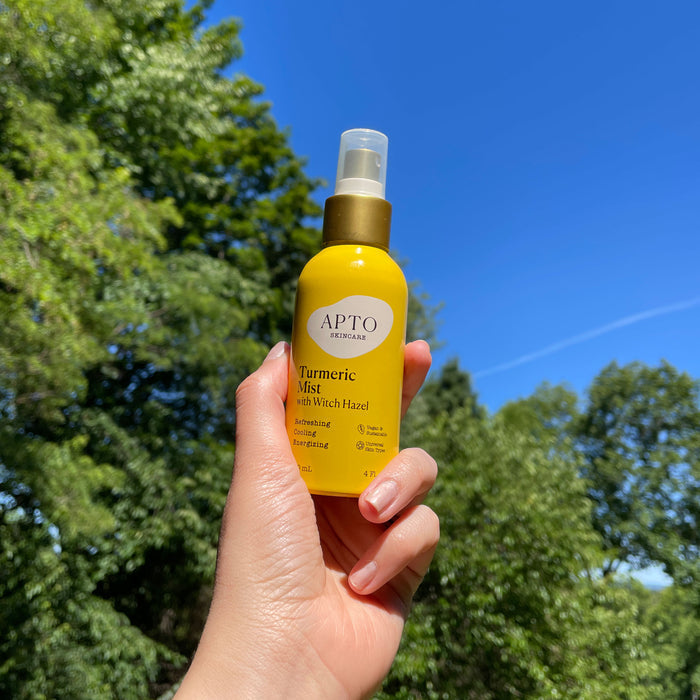 A hand holds APTO Skincares Turmeric Mist with Witch Hazel, Reviving & Brightening Facial Spritz against a clear blue sky and green trees, promising glowing skin with every cruelty-free spray.