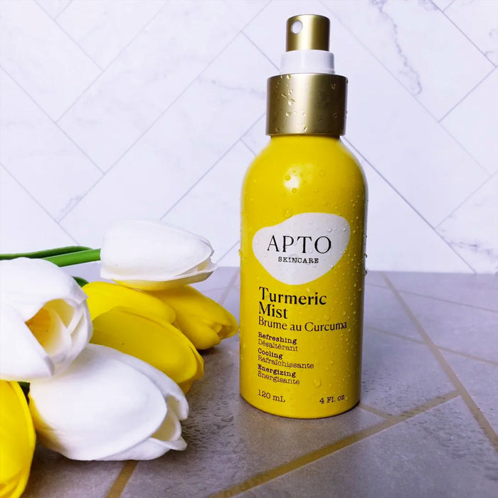 A yellow spray bottle, APTO Skincare Turmeric Mist with Witch Hazel, sits on tiles, promising glowing skin with its refreshing, hydrating benefits and cruelty-free formula. Yellow and white tulips beside it add a contrasting touch to the scene.