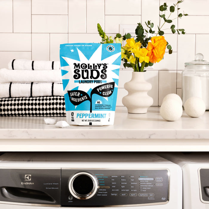 A bag of Molly's Suds Ultra Concentrated Laundry Detergent Pods rests on a laundry room counter next to folded towels, a white vase with flowers, and wool dryer balls. A washing machine sits below—ideal for sensitive skin.