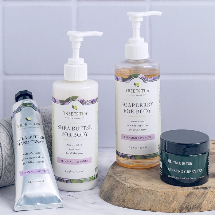 Displayed on a light surface is a collection of Tree To Tub skincare products, including Ultra Hydrating Body Wash for Dry Skin, shea butter hand cream and body lotion, soapberry hydrating body wash, and ginseng green tea face cream, all in pastel packaging with lavender accents.