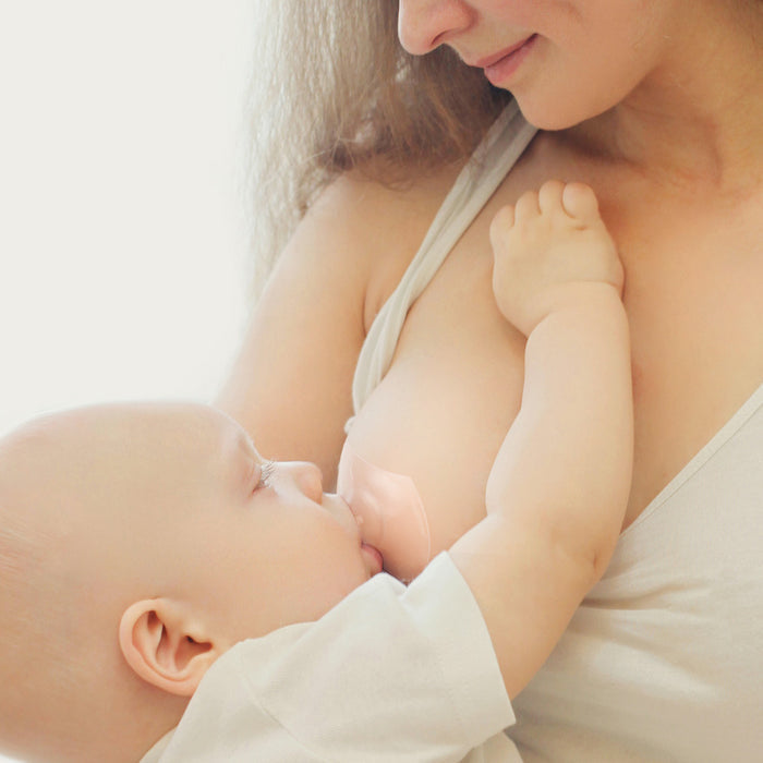 A mother uses Haakaa Ultra-Thin Silicone Nipple Shields, Size M (18mm), to protect her sore nipples while breastfeeding her baby. The baby nurses contentedly as she gazes affectionately at them, their bond tenderly highlighted by the soft lighting.