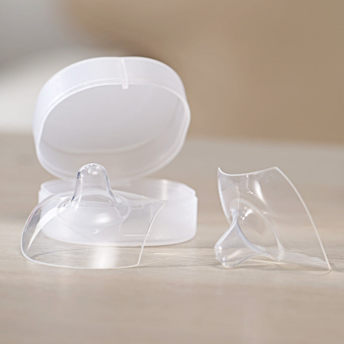 Two Haakaa Ultra-Thin Silicone Nipple Shields, Size M (18mm), are shown on a wooden surface. One shield is in an open plastic case, while the other rests adjacent. A softly blurred background highlights these breastfeeding essentials for soothing sore nipples.