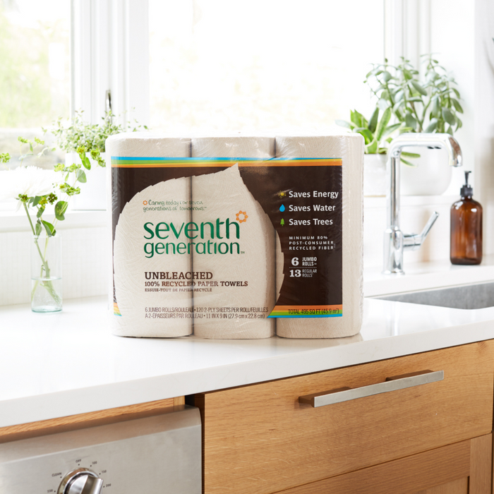 A pack of Seventh Generation Unbleached Paper Towels (120 sheets per roll), made from recycled paper and eco-friendly packaging, is on a kitchen counter by a window, with bottles and a potted plant in the background.