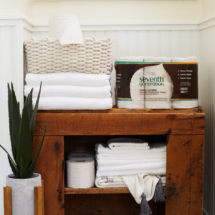 A wooden shelf displaying white towels holds a basket with Unbleached Paper Towels (120 sheets per roll) and Seventh Generation toilet tissue, alongside an eco-friendly potted plant, set against a white paneled wall.