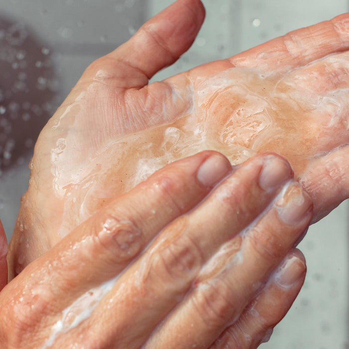 A person washes hands with soap and water using OSEAs Undaria Cleansing Body Polish, featuring AHA exfoliation. Bubbles and water droplets highlight cleanliness and rejuvenation.