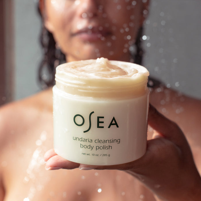 In a softly blurred bathroom setting, someone holds a jar of OSEAs Undaria Cleansing Body Polish, recognized for its creamy gel-to-milk texture. The label is visible, highlighting its enriching Undaria seaweed and AHA exfoliation properties.