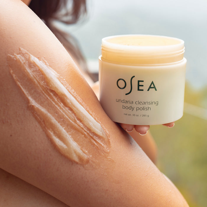 Close-up of a persons arm with a smear of gel-to-milk body polish. They hold an Undaria Cleansing Body Polish by OSEA, featuring undaria seaweed and AHA exfoliation. The blurred background emphasizes the products transformative skin effect.