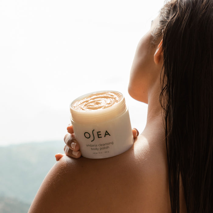 A person with wet hair holds a large container of OSEAs Undaria Cleansing Body Polish against their shoulder, set against a blurred natural backdrop. This gel-to-milk formula, enriched with Undaria seaweed and AHA, gently exfoliates for rejuvenated skin.