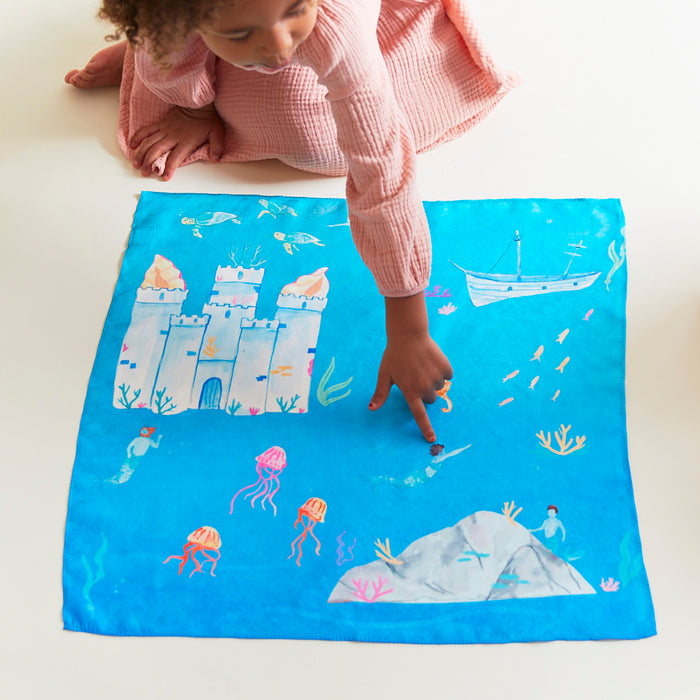 A child in a pink dress excitedly points at the Under the Sea Play Map by Sarahs Silks, a vibrant blue mat made with non-toxic dyes depicting an underwater scene with a castle, jellyfish, boat, and fish.