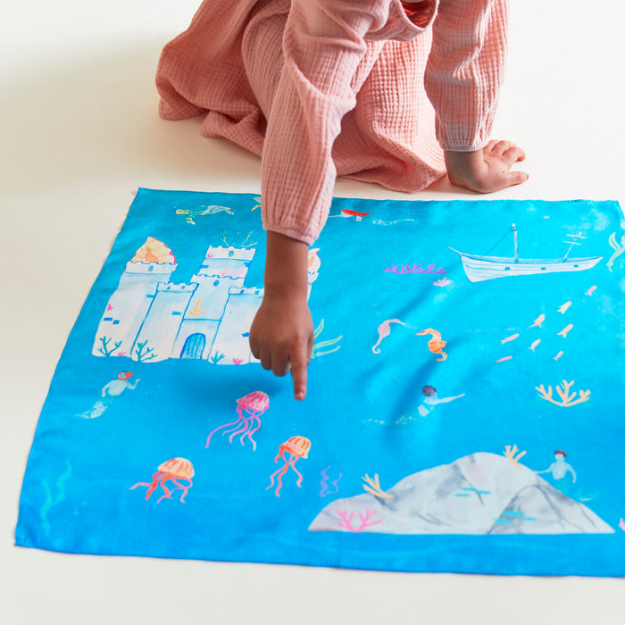 A child in a pink outfit points at Sarahs Silks Under the Sea Play Map, featuring an underwater theme with a castle, jellyfish, sea creatures, and coral. Crafted with non-toxic dyes, this vibrant mat is laid out invitingly on a light surface.