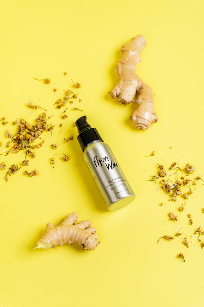 A silver spray bottle with a black cap sits on a vibrant yellow background, surrounded by ginger pieces and dried flowers. The label reads Underarm Oil by PiperWai, celebrated for its natural deodorant properties boosted by essential oils.