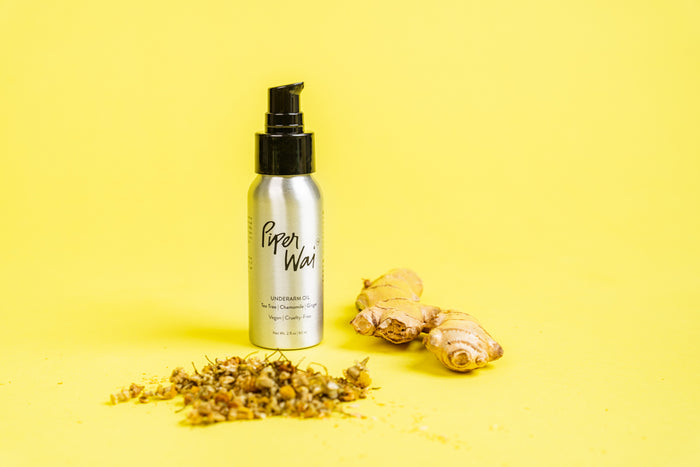A silver bottle with a black pump, labeled Underarm Oil from PiperWai, sits proudly on a bright yellow background. This natural deodorant is paired with small pieces of ginger root and dried herbs, emphasizing its essential oil blend for a refreshing touch.