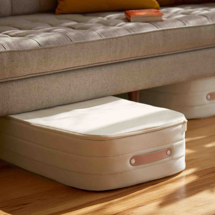 An Open Spaces beige Underbed Storage bag made of natural canvas with breathable protection is under a gray sofa on a wooden floor, next to an identical bag. A small orange book rests on the sofa.
