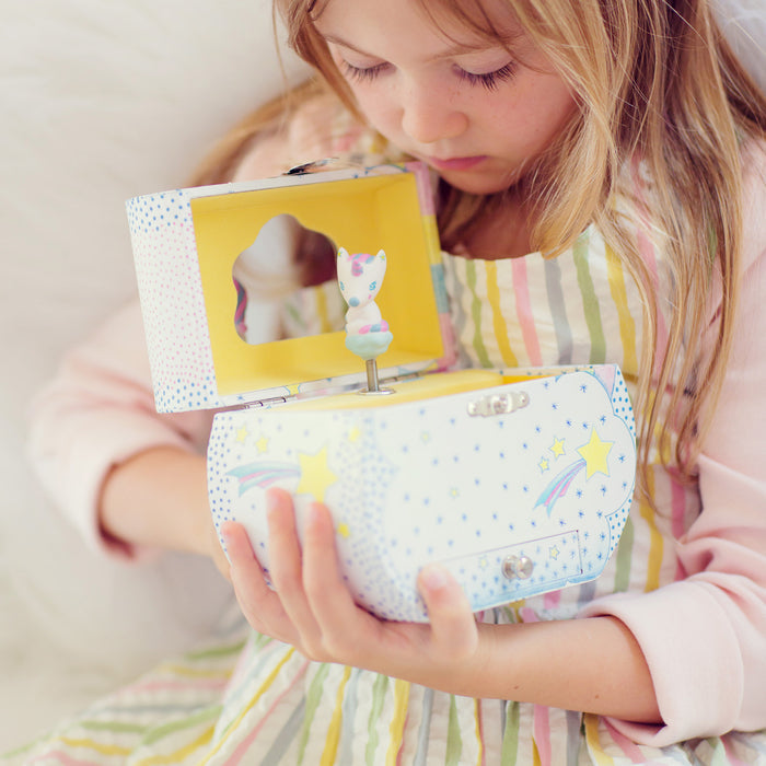 A young girl, wearing a pastel-striped dress and seated on a soft surface, admires the DJECO Unicorn Dreams Musical Treasure Box. This enchanting jewelry storage is adorned with stars and rainbows and features a twirling unicorn, captivating her in its magical allure.