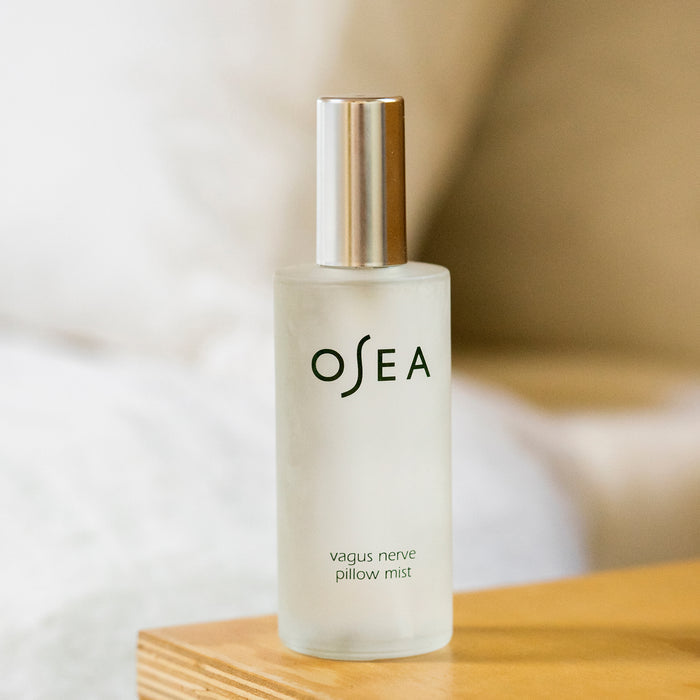 A frosted glass bottle of OSEA Vagus Nerve Pillow Mist with a silver cap sits on a wooden surface, evoking relaxation, while a soft-focus bed and pillows appear in the background.
