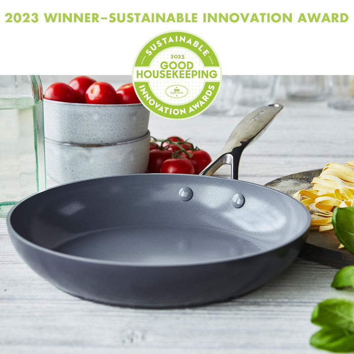 A non-stick frying pan sits on a wooden surface with tomatoes, pasta, and a basil plant around it. The image includes a 2023 Winner - Sustainable Innovation Award badge from Good Housekeeping at the top.