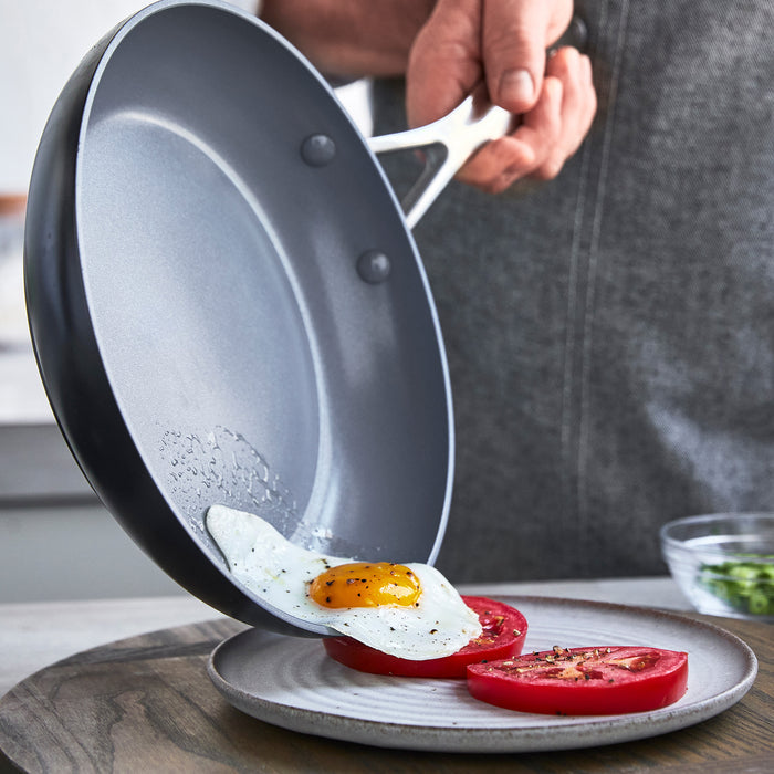 A person gently slides a sunny-side-up egg from a GreenPan Valencia Pro Ceramic, Nonstick, Hard Anodized 10 Fry Pan onto a plate of sliced tomatoes. The softly blurred kitchen countertop in the background creates an inviting atmosphere for culinary creativity.
