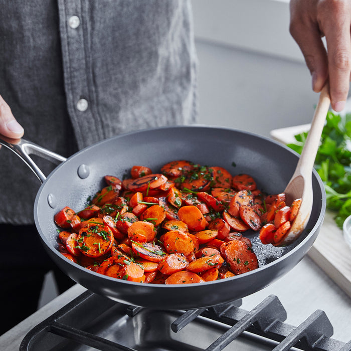 Someone is using a GreenPan Valencia Pro Ceramic, Nonstick, Hard Anodized Fry Pan to cook sliced carrots on a stove. The PFAS-Free cookware allows for safe and healthy cooking as the seasoned carrots are stirred with a wooden spoon. A cutting board with parsley is seen in the background.
