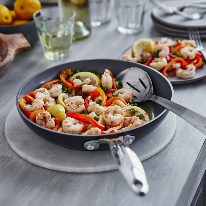 A Valencia Pro Ceramic Nonstick 10 Fry Pan by GreenPan, used to prepare shrimp with bell peppers and lemon wedges, sits on a gray table. A serving spoon is inside, while plates of the meal and a glass of white wine are seen in the background. The cookware is PFAS-free for healthy meals.