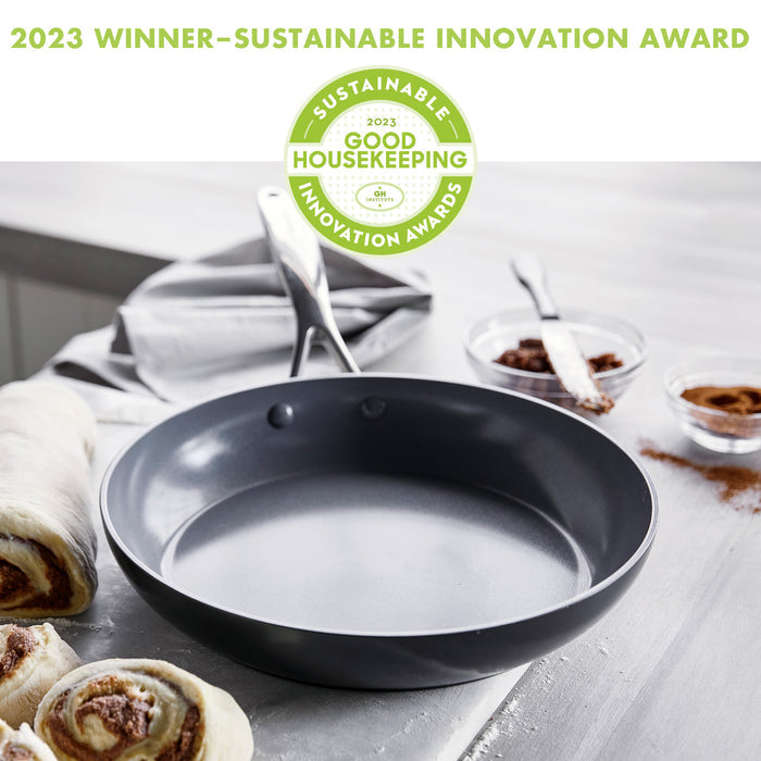 A non-stick frying pan on a kitchen counter, surrounded by baking ingredients like cinnamon rolls and bowls of cinnamon. "2023 Winner–Sustainable Innovation Award" and "Good Housekeeping Sustainable Innovation Awards" badges are displayed above the pan.
