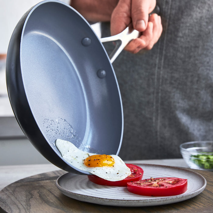 A hand tilts the GreenPan Valencia Pro Ceramic Nonstick 12 Fry Pan, sliding a sunny-side-up egg onto a plate with sliced tomatoes. The kitchen background is blurred, emphasizing the PFAS-free ceramic nonstick coating for healthier cooking.