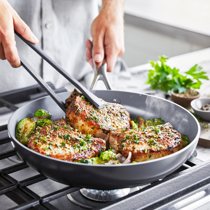 Using tongs, a person cooks seasoned pork chops in a GreenPan Valencia Pro Ceramic Nonstick 12 Fry Pan on the stove. The hard anodized aluminum ensures even heating, while the PFAS-free design keeps it safe. Herbs garnish the meat alongside colorful veggies, and fresh parsley and spices are visible nearby.