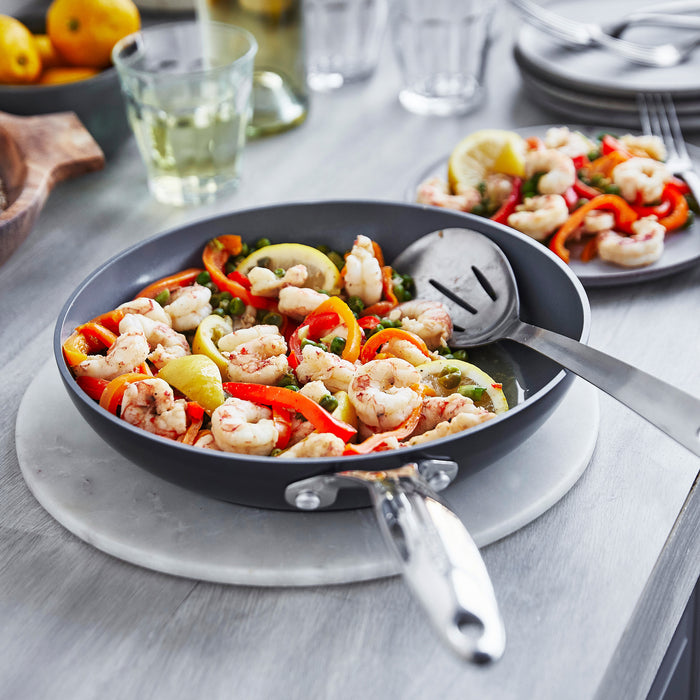 A GreenPan Valencia Pro Ceramic Nonstick 12 Fry Pan brimming with shrimp, red bell peppers, and green veggies sits on a gray marble trivet. In the background, similar dishes and a glass of white wine adorn a wooden table.